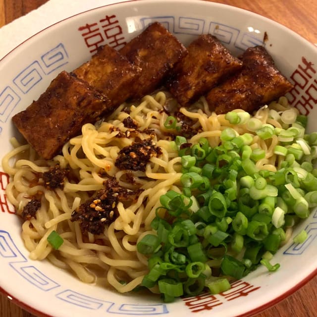 A bowl of noodles topped with a handful of green onions and strips of sticky red tofu