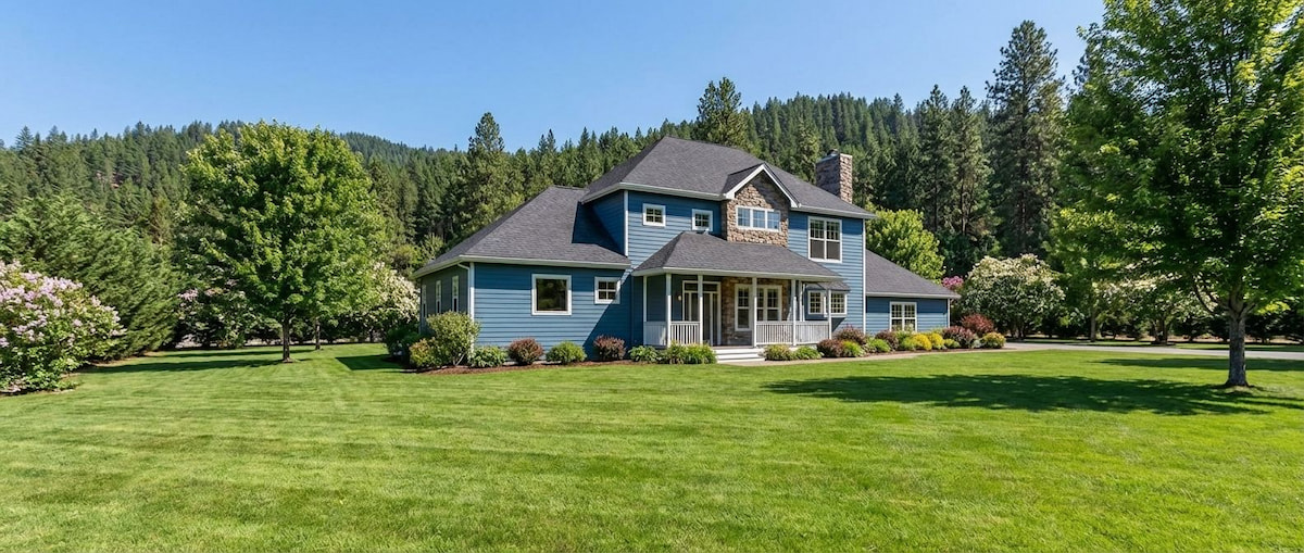 Large blue two-story house featuring multiple roof gables, a covered front porch with white railings, and surrounded by a spacious lawn with wooded background.