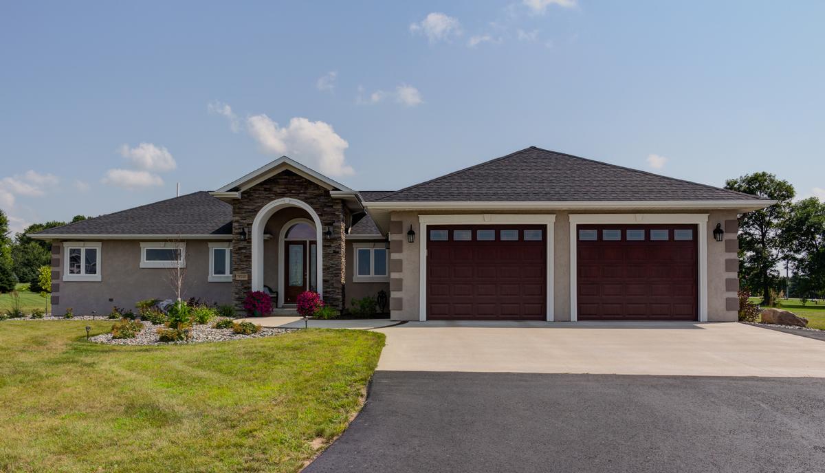 Stucco-finished modular home by Wisconsin Homes with stone arched entryway, dark wood garage doors, and a landscaped front yard.