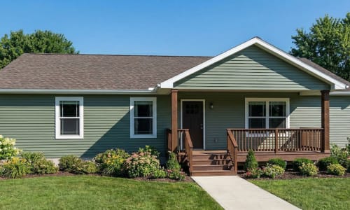 Green cottage-style modular home by Wisconsin Homes in Marshfield, WI with covered front porch and landscaped yard.
