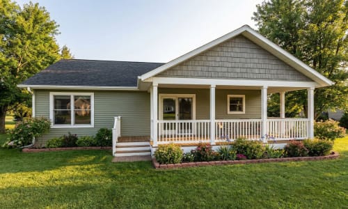 Green modular home with wide front porch, white railings, and flower garden by Wisconsin Homes in Marshfield, WI.