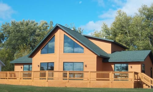 Log cabin-style chalet modular home by Wisconsin Homes with expansive windows, green shingle roof, and a full wraparound wooden deck surrounded by trees.