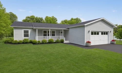 White single-story ranch-style home with black roof, attached one-car garage, covered front porch, and neatly trimmed shrubs on a large green lawn.