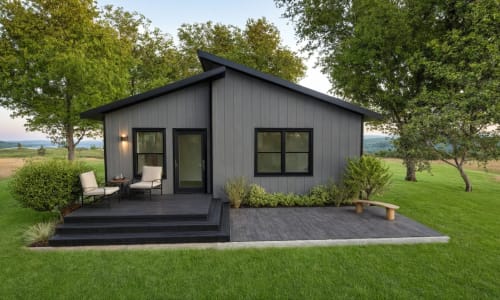 Minimalist dark-gray cabin with a front deck, black-trimmed windows, outdoor seating, and surrounded by a green lawn and open landscape.