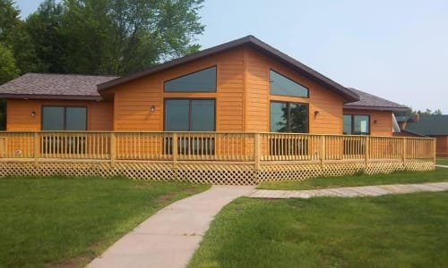 Cedar cottage-style home by Wisconsin Homes with a wraparound wood deck, large windows, and manicured lawn.