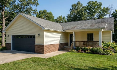 Yellow modular home with dark garage door, brick skirt, covered porch, and flower garden by Wisconsin Homes in Marshfield, WI.