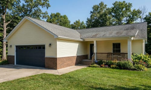 Yellow modular home with dark garage door, brick skirt, covered porch, and flower garden by Wisconsin Homes in Marshfield, WI.