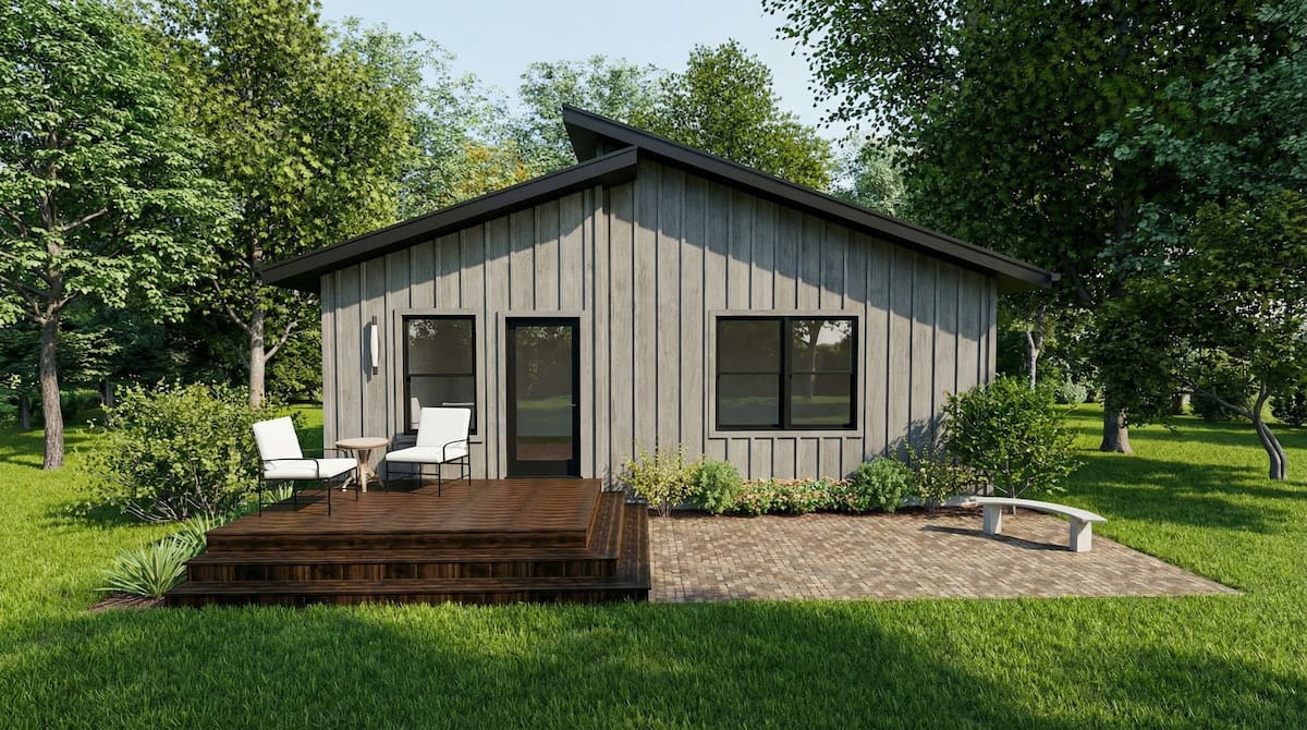 Minimalist dark-gray cabin with a front deck, black-trimmed windows, outdoor seating, and surrounded by a green lawn and open landscape.
