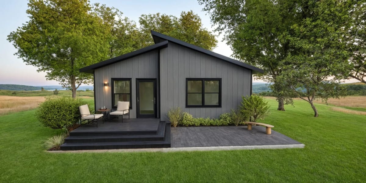 Minimalist dark-gray cabin with a front deck, black-trimmed windows, outdoor seating, and surrounded by a green lawn and open landscape.