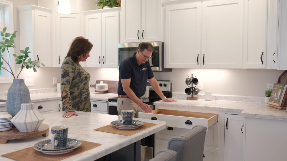 Wisconsin Homes representative showing kitchen drawer features in model home with white cabinets and marble countertops.