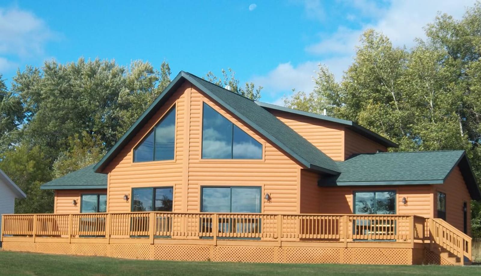 Log cabin-style chalet modular home by Wisconsin Homes with expansive windows, green shingle roof, and a full wraparound wooden deck surrounded by trees.