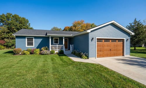 Blue modular home with wood-tone garage door, covered front entry, and manicured landscaping by Wisconsin Homes in Marshfield, WI.