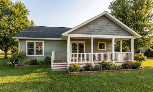 Green modular home with wide front porch, white railings, and flower garden by Wisconsin Homes in Marshfield, WI.