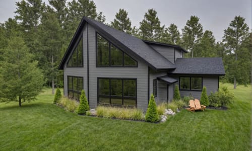 Modern black chalet-style home with sharp rooflines and large floor-to-ceiling windows, surrounded by landscaped greenery and forested background.