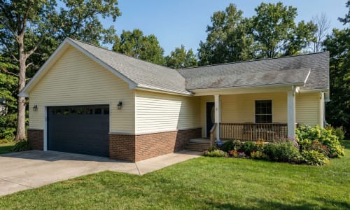 Yellow modular home with dark garage door, brick skirt, covered porch, and flower garden by Wisconsin Homes in Marshfield, WI.