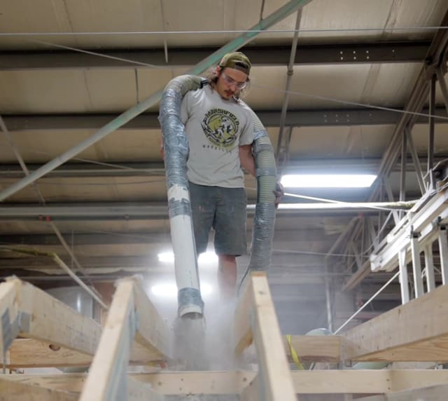 Wisconsin Homes worker installing insulation into floor system at factory using large hose equipment.