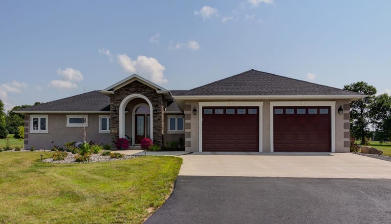 Stucco-finished modular home by Wisconsin Homes with stone arched entryway, dark wood garage doors, and a landscaped front yard.