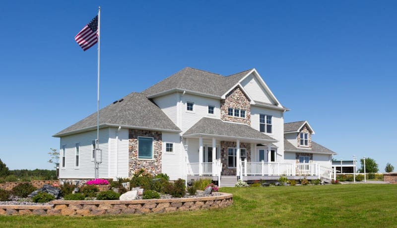 Two-story white modular home by Wisconsin Homes featuring stone accents, a large front porch with white railing, gray roof, and manicured landscaping under a clear blue sky.