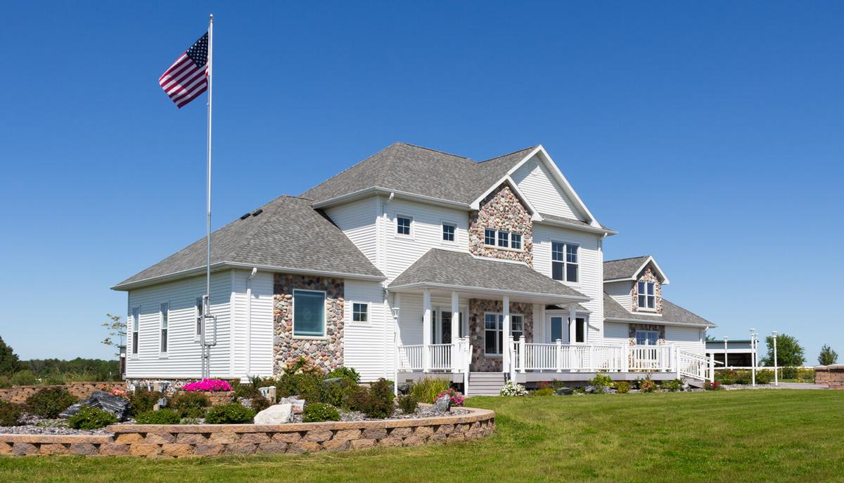 Two-story white modular home by Wisconsin Homes featuring stone accents, a large front porch with white railing, gray roof, and manicured landscaping under a clear blue sky.