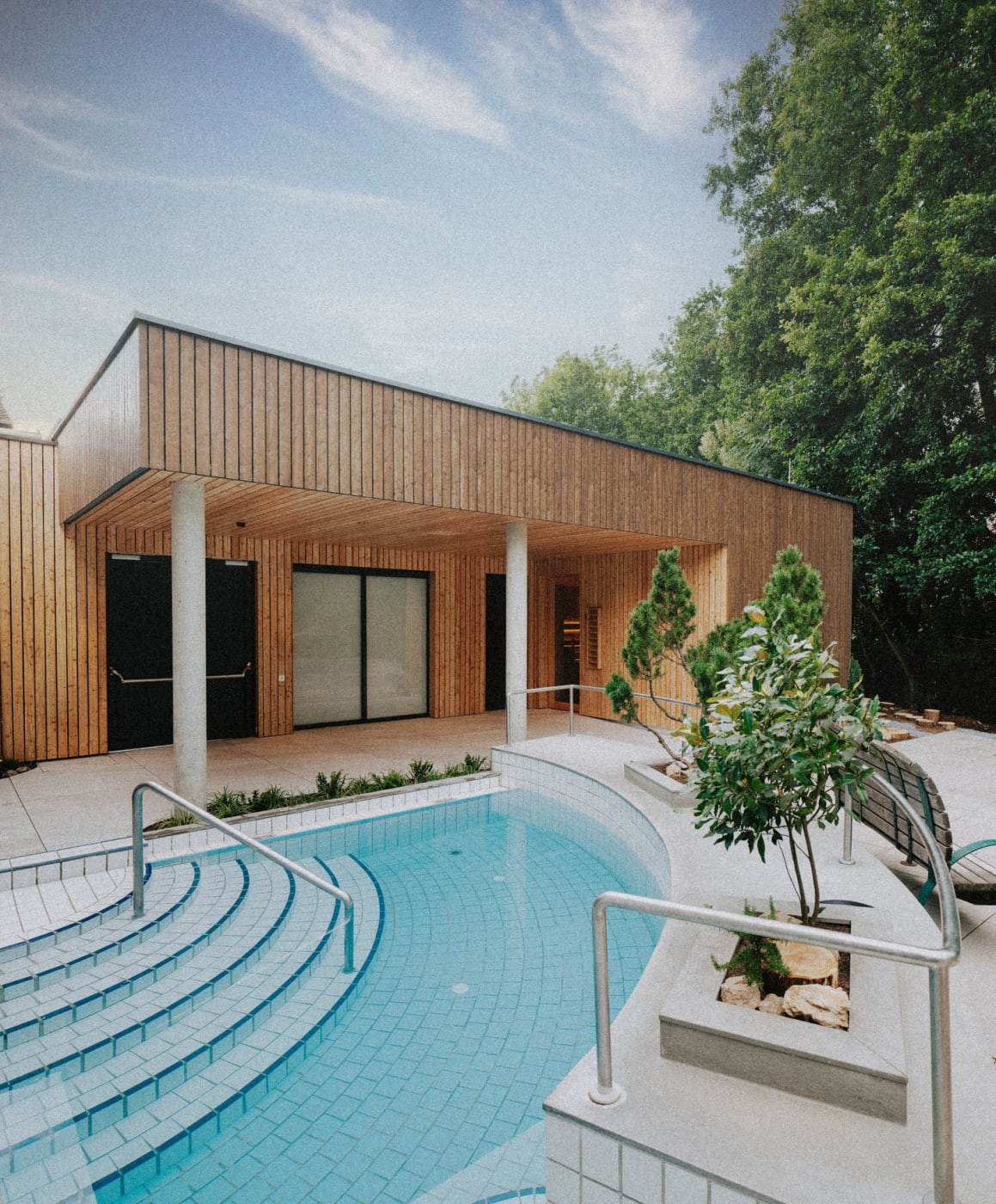 O pavilhão de madeira da Quell-Sauna acima de uma piscina externa cercada por árvores