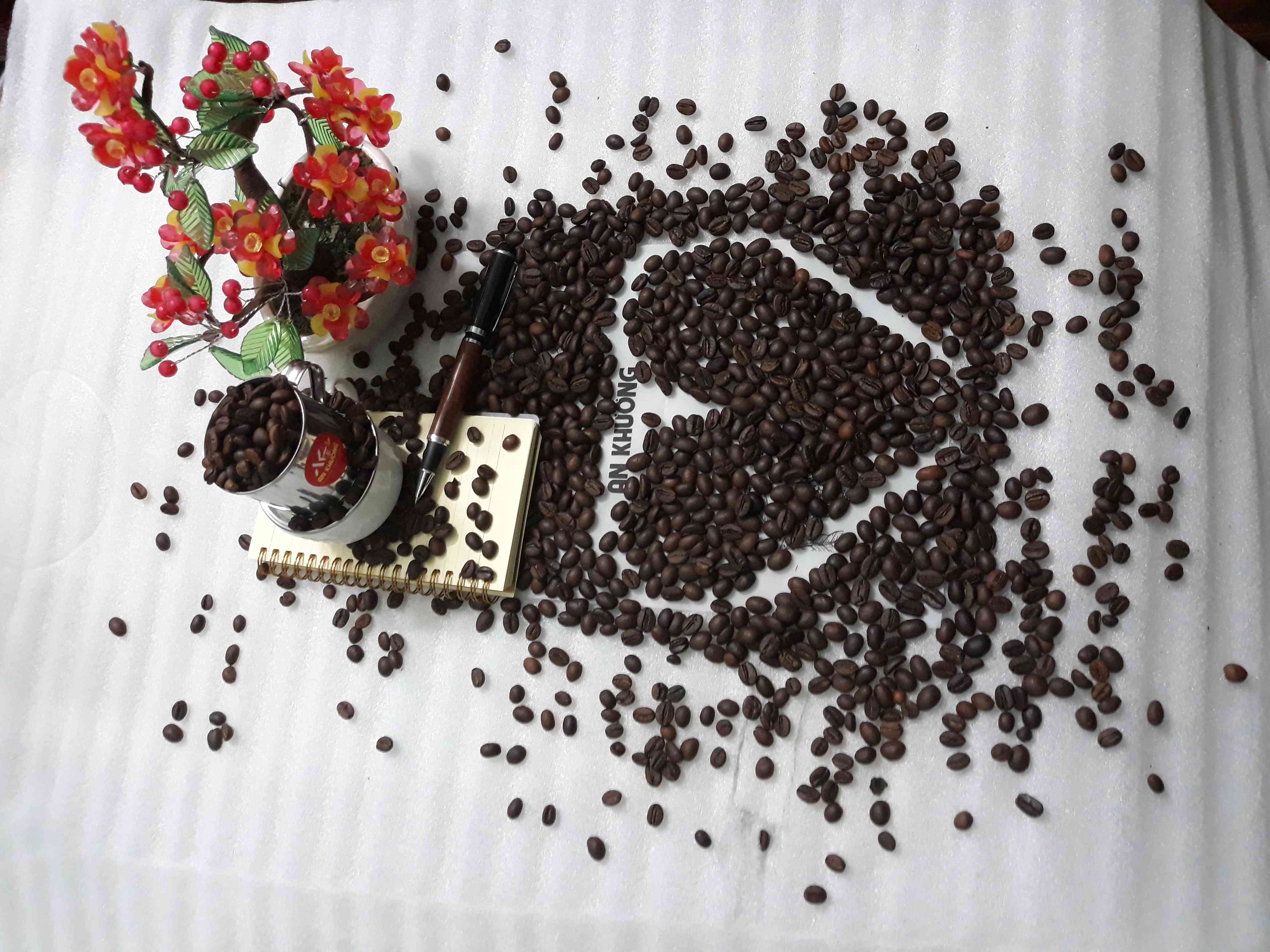 Flatlay hạt cà phê tạo hình logo/chữ An Khương, kèm hoa, bút, sổ và phin