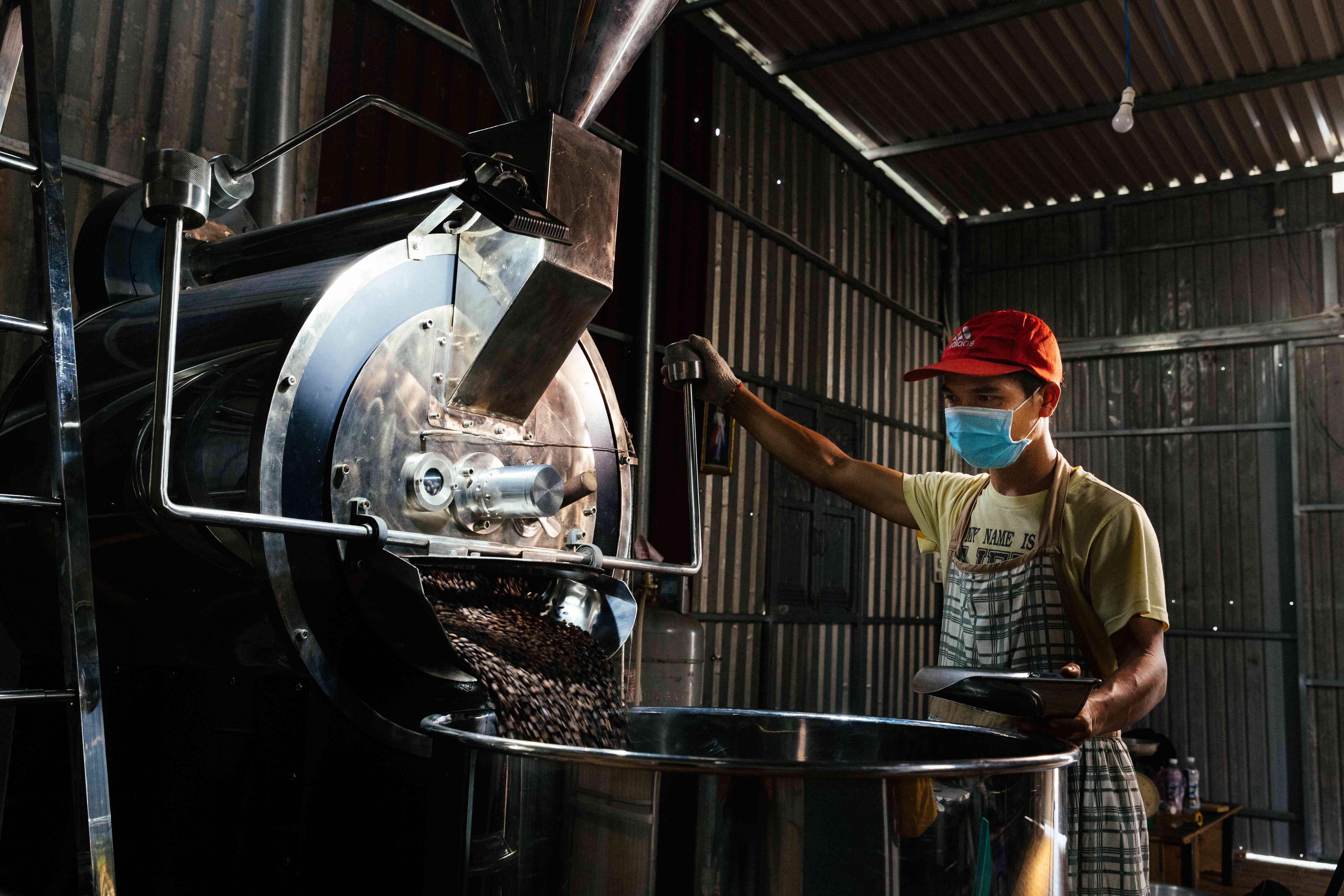 Toàn cảnh máy rang công nghiệp trong xưởng, công nhân đứng vận hành và hạt chảy xuống khay.