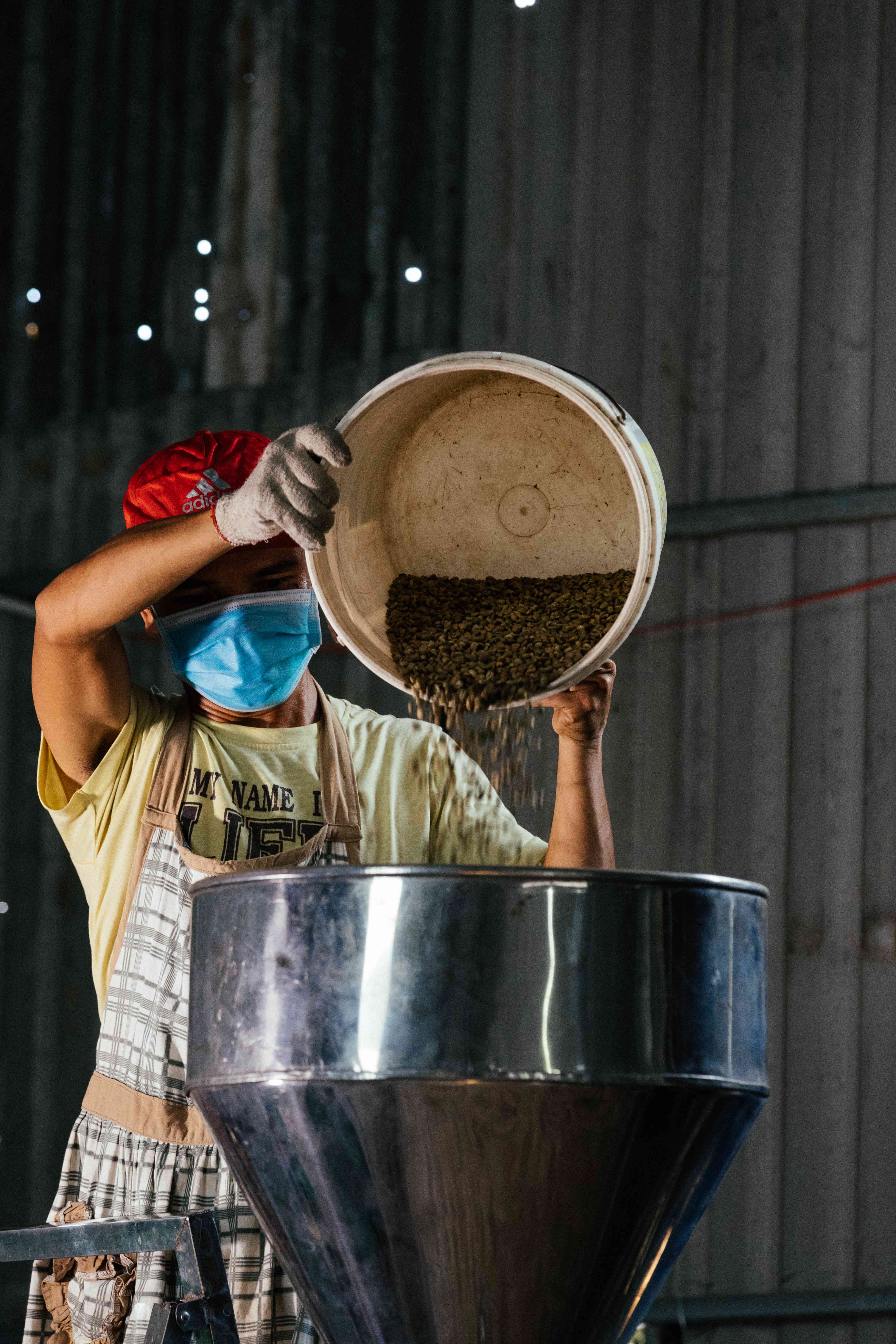 Người công nhân đổ hạt từ thùng nhựa lớn vào phễu/máy, đang đeo khẩu trang và tạp dề.
