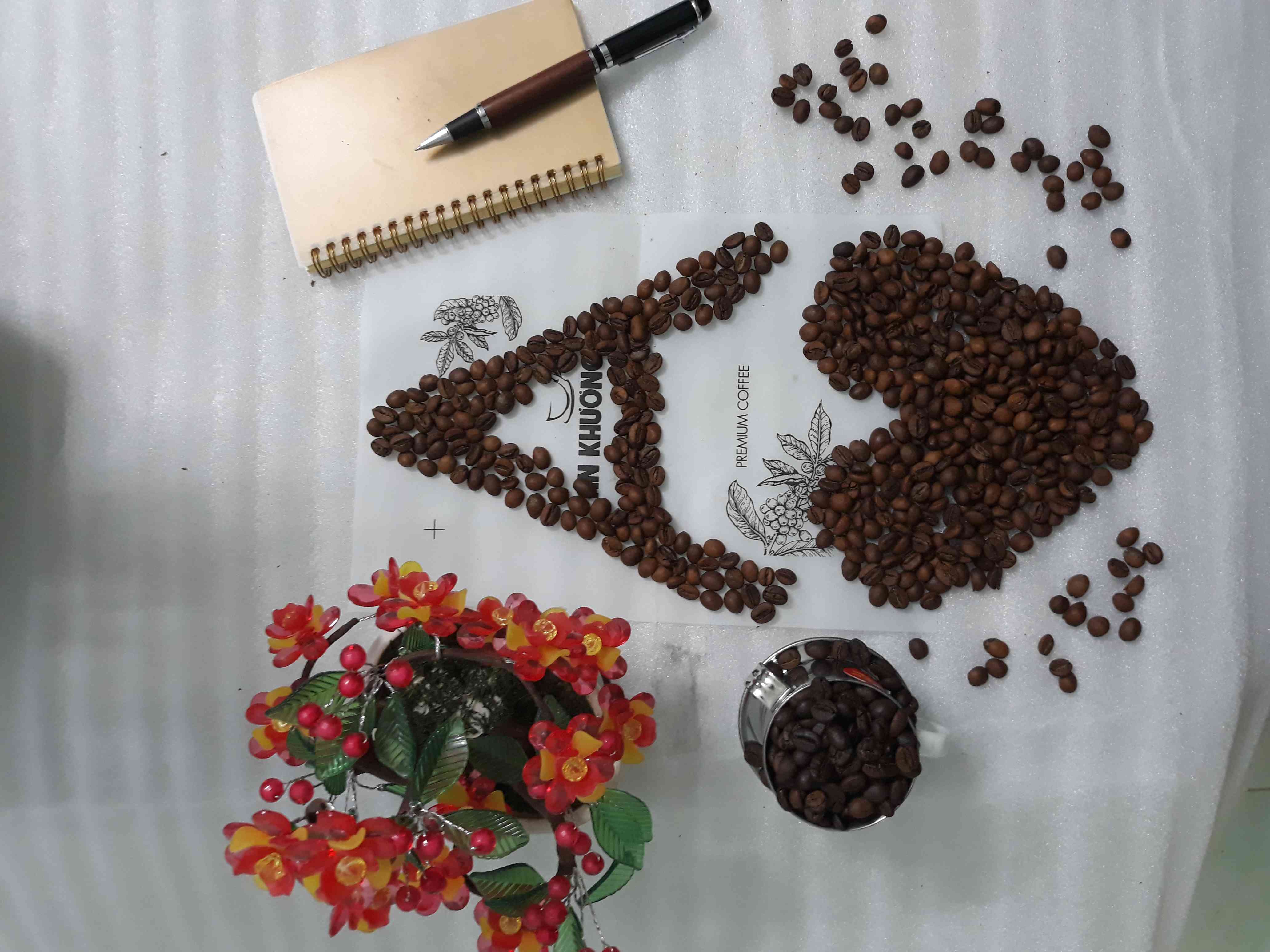 Hạt cà phê được xếp thành hình minh họa trên bao/tấm có chữ AN KHƯƠNG và dụng cụ ghi chép, hoa trang trí.