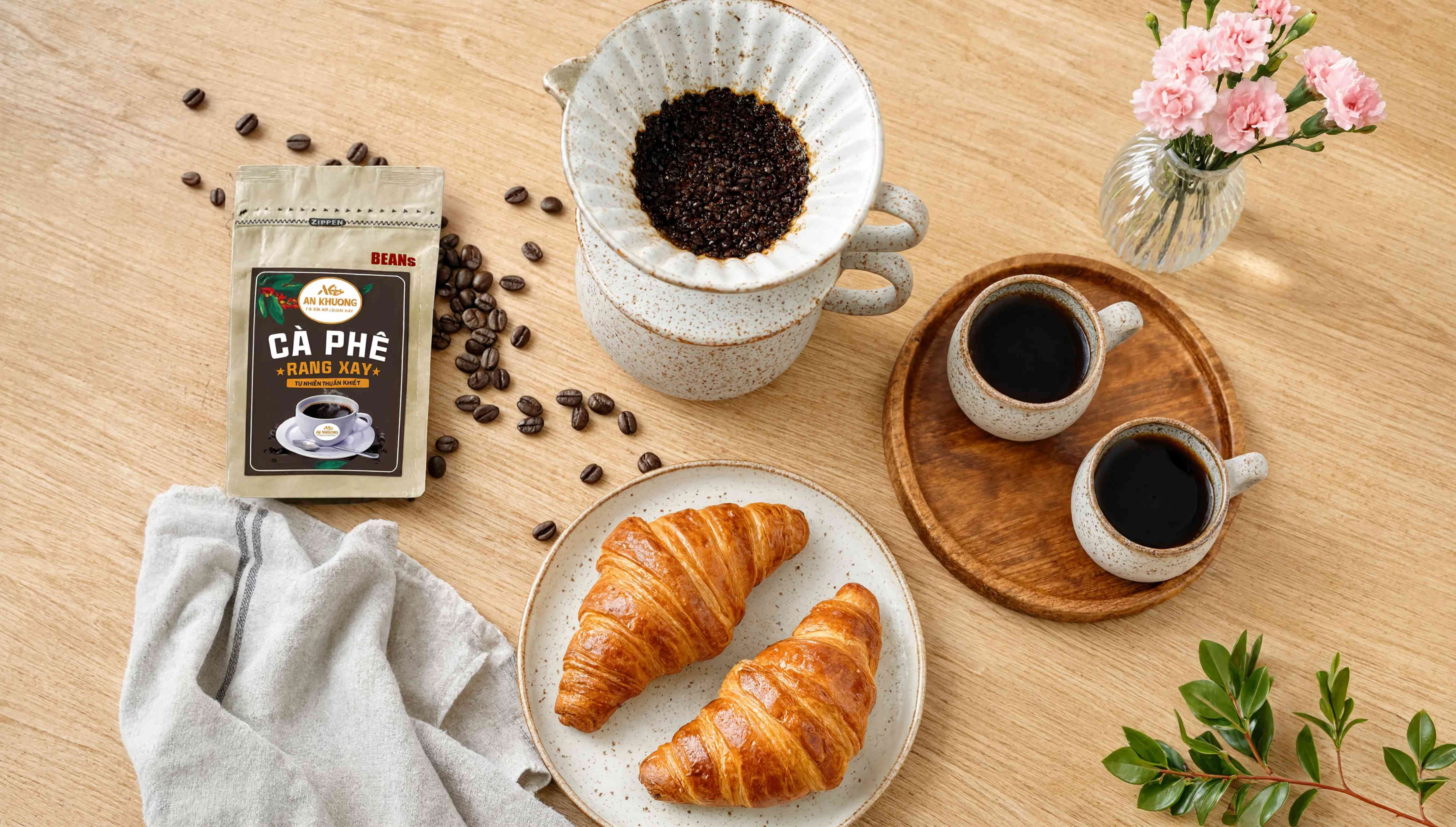 Flatlay bàn ăn sáng có túi cà phê An Khương, phin, hai ly cà phê, croissant và hoa