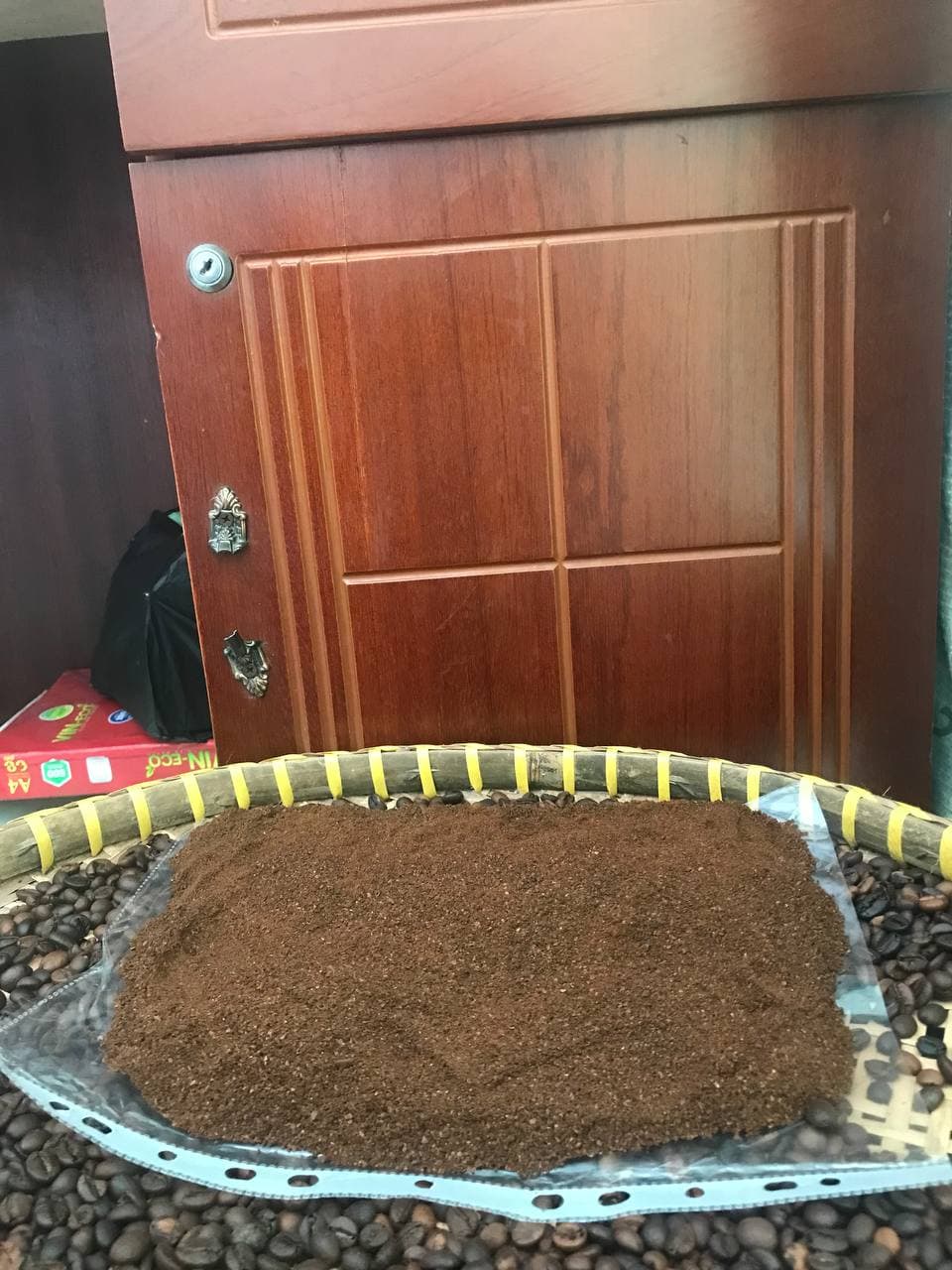Bột cà phê được dàn thành mảng lớn trên khay, bao quanh bởi hạt rang; có cửa gỗ phía sau