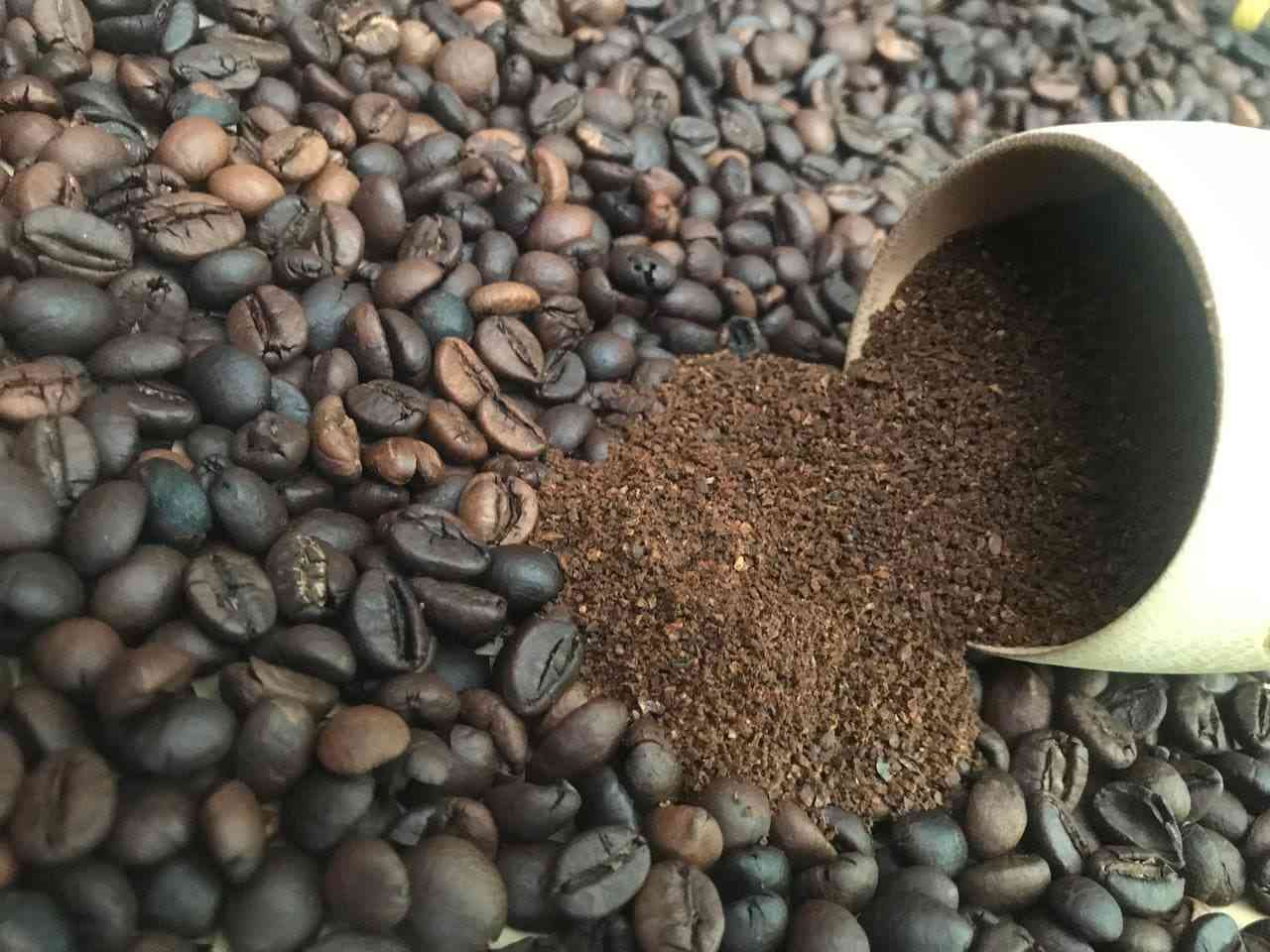 Cốc/ống nghiêng đổ bột cà phê xay lên nền nhiều hạt rang, nhìn cận.