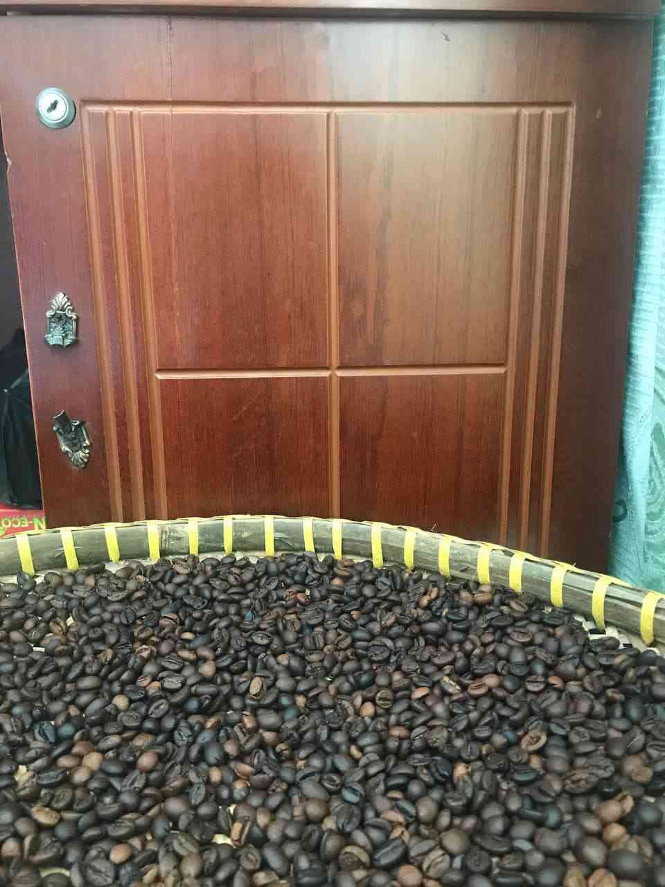 Mẹt lớn đầy hạt cà phê rang, góc chụp rộng hơn; cửa gỗ và rèm xanh ở phía sau