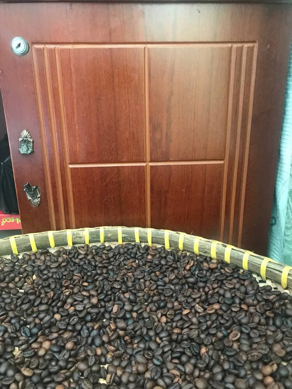 Cùng cảnh mẹt đầy hạt cà phê rang, chụp ngang; hạt chiếm nửa dưới và cửa gỗ ở hậu cảnh