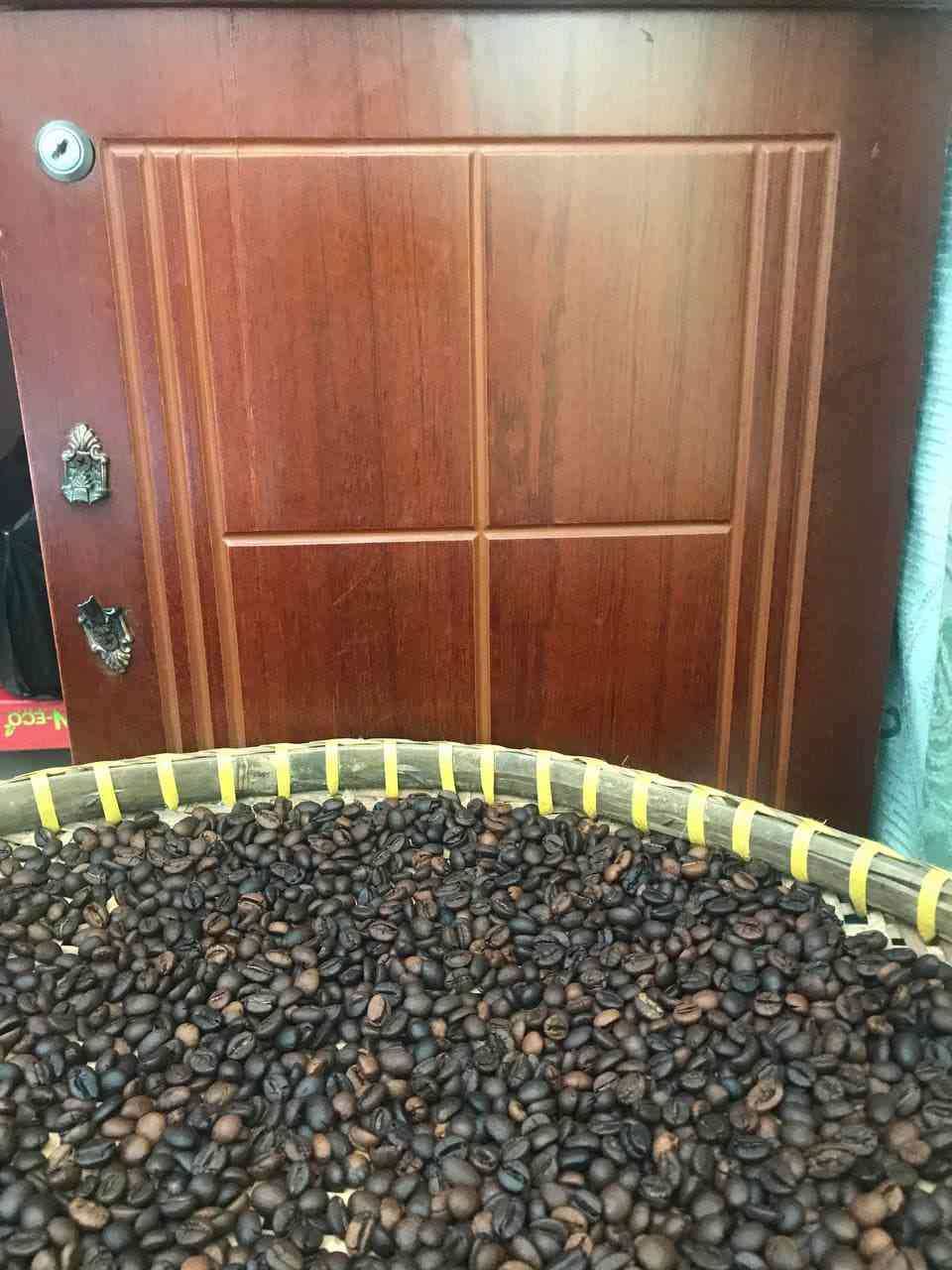 Mẹt lớn chứa lượng hạt cà phê rang rất nhiều, chụp từ phía trước; nền là cánh cửa gỗ
