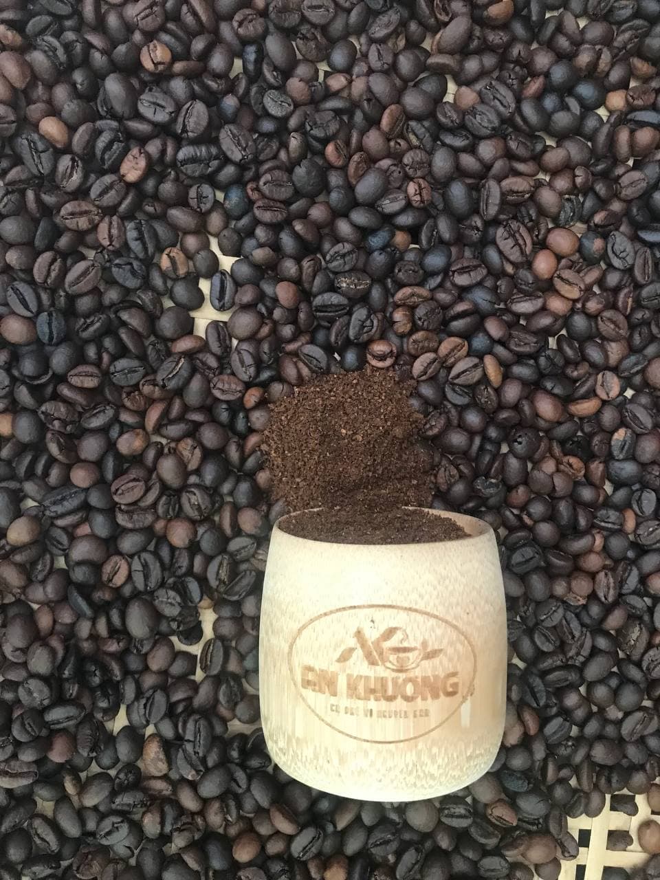 Ly tre An Khương trên nền hạt cà phê rang, bột cà phê rơi vào ly; bố cục dọc, chủ thể ở trung tâm