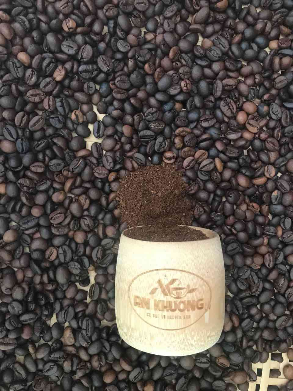 Ly tre An Khương đặt giữa nền hạt cà phê rang; bột cà phê được đổ/tạo thành dòng phía trên miệng ly