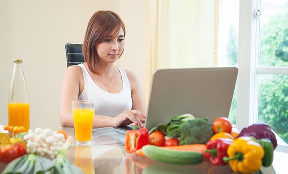Wanita menggunakan laptop diatas meja disamping jus jeruk dan paprika, timun, dan tomat