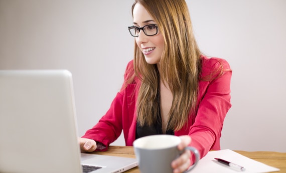 Wanita menggunakan jas pink sedang menggunakan laptop sambil memegang cangkir kopi