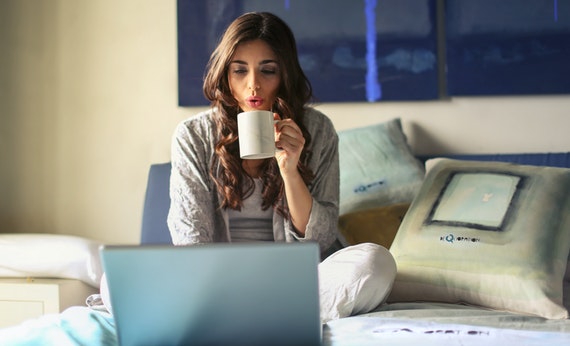 Wanita sedang minum kopi diatas kasur sambil menggunakan laptop