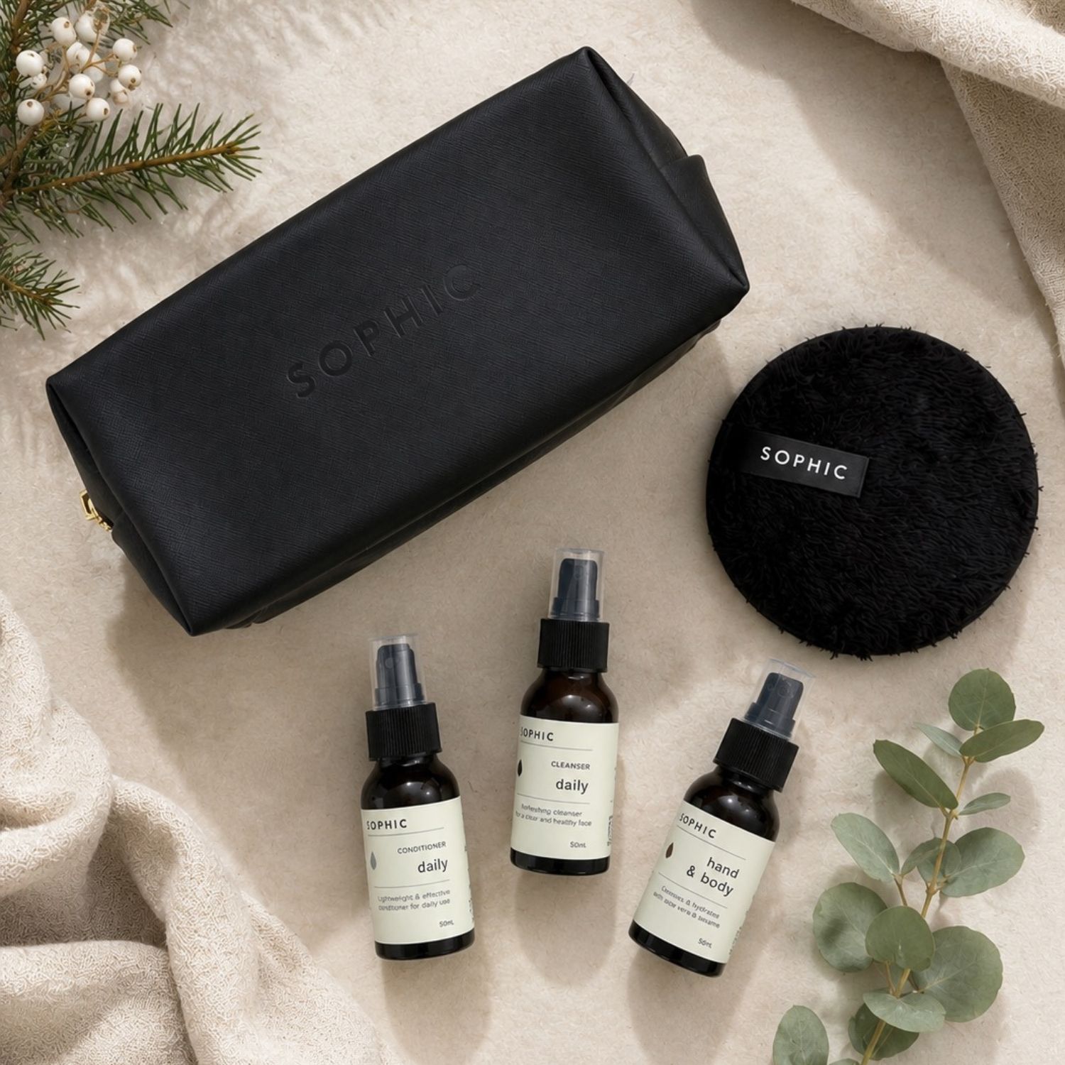 Sophic black embossed cosmetic bag with three small amber bottles and a black cleansing pad on a cream blanket