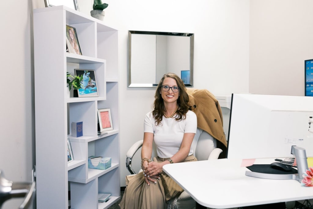 A smiling professional woman sitting at a white desk with a desktop computer inside a private office suite, featuring a modern white geometric bookshelf with framed photos and a square wall mirror.
