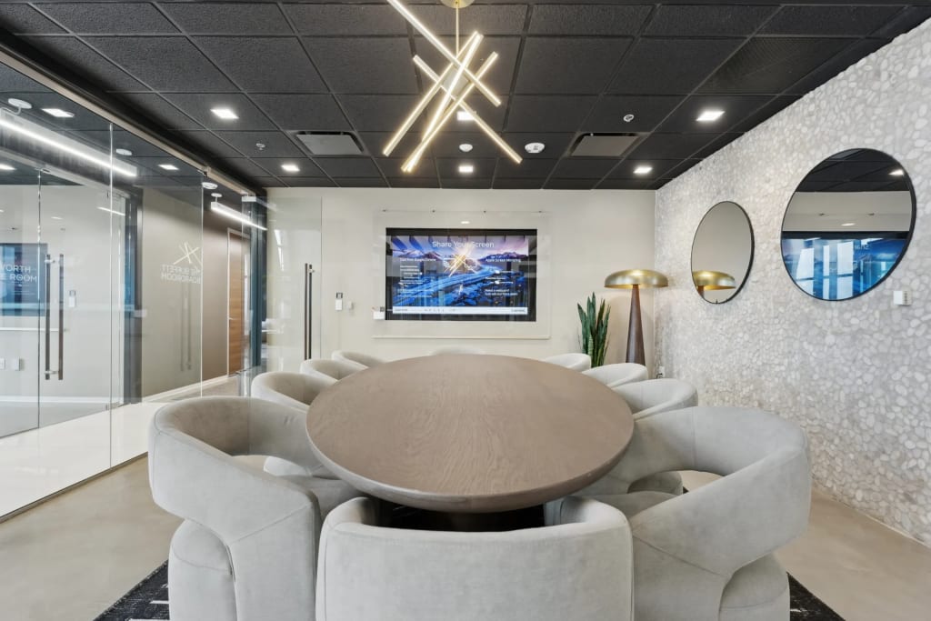 A luxury executive boardroom at Lucid Private Office featuring a large oval wooden conference table, plush grey fabric chairs, round mirrors on a textured pebble wall, and a modern geometric gold chandelier.