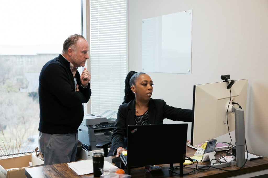 A man reviewing work over a colleague's shoulder at a desk inside a private office, featuring a desktop monitor, a laptop, a window with blinds, and a blank glass whiteboard on the wall.