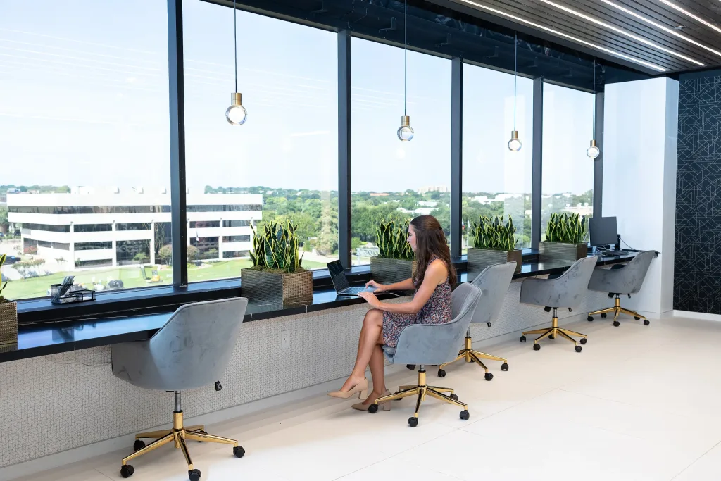 A woman working on a laptop at a long window-side coworking desk inside a Lucid Private Office Las Colinas location, featuring plush grey swivel chairs with gold bases, modern pendant lighting, snake plants in decorative planters, and floor-to-ceiling windows overlooking a suburban landscape.