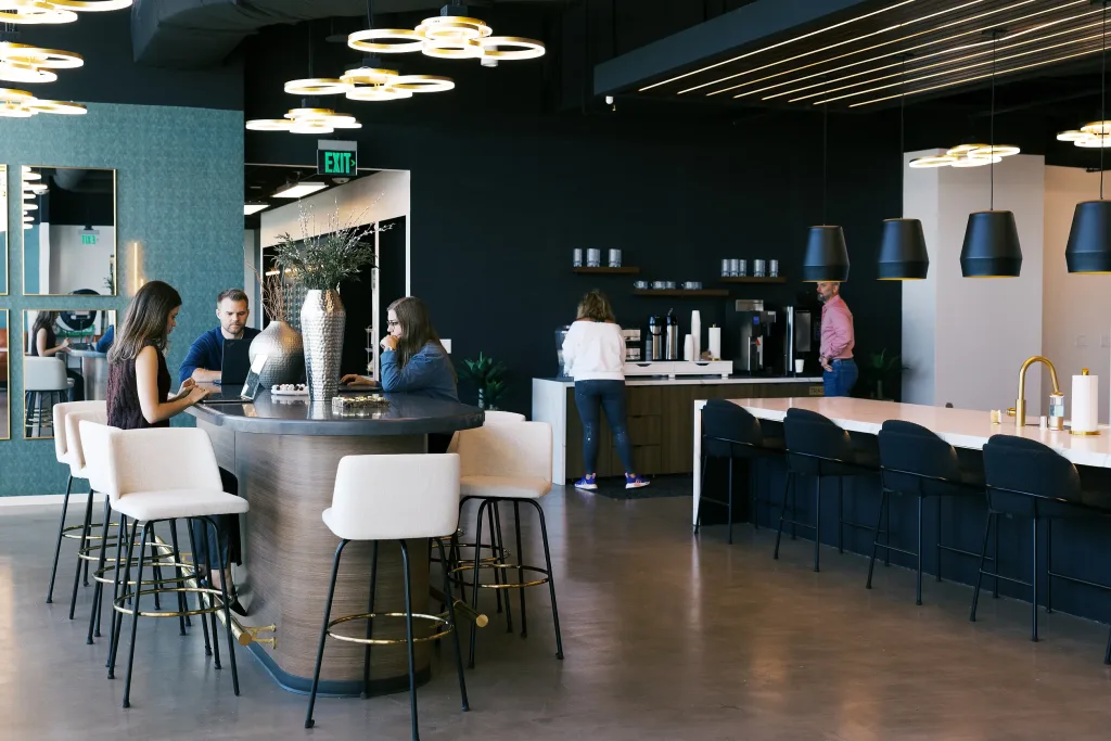 Corporate breakroom and coworking cafe at Lucid Private Office, showing professionals working at a high-top island with white barstools, a fully equipped coffee bar, and modern geometric halo lighting fixtures.