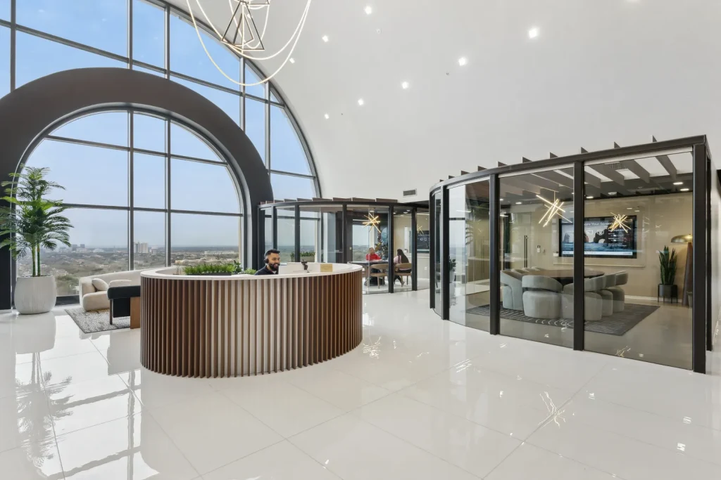 Bright, high-ceiling reception lobby at Lucid Private Office featuring a curved wood-slat front desk, polished white tile floors, a massive arched window with city views, and glass-walled private conference rooms.