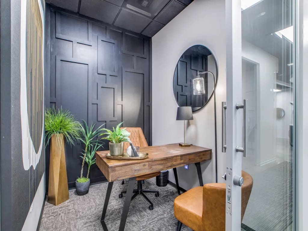 A compact private office suite at Lucid Private Office showcasing a wooden desk, tan leather chairs, a round wall mirror, a sleek task lamp, and a dark geometric paneled accent wall.