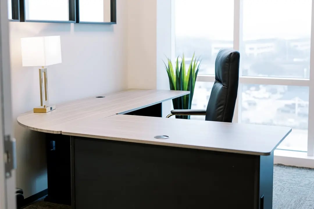 Close-up of a modern office desk with a black executive chair and a tall plant by the window