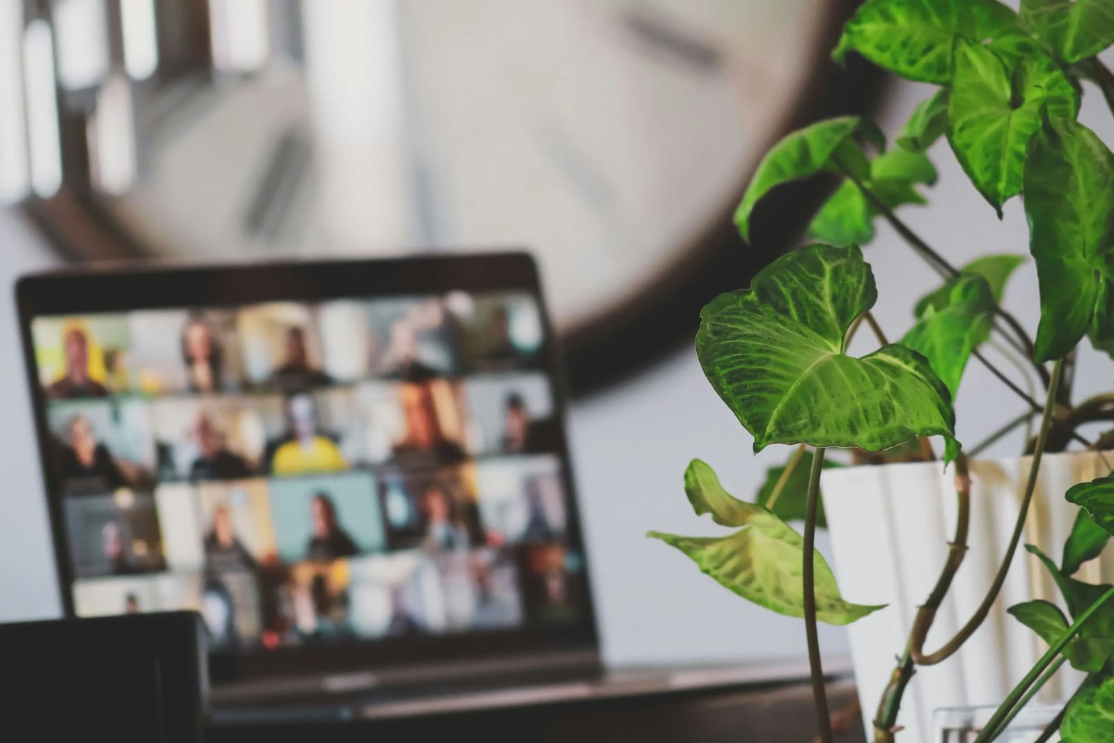 Laptop displaying a large video conference call with a plant and clock in a blurred background