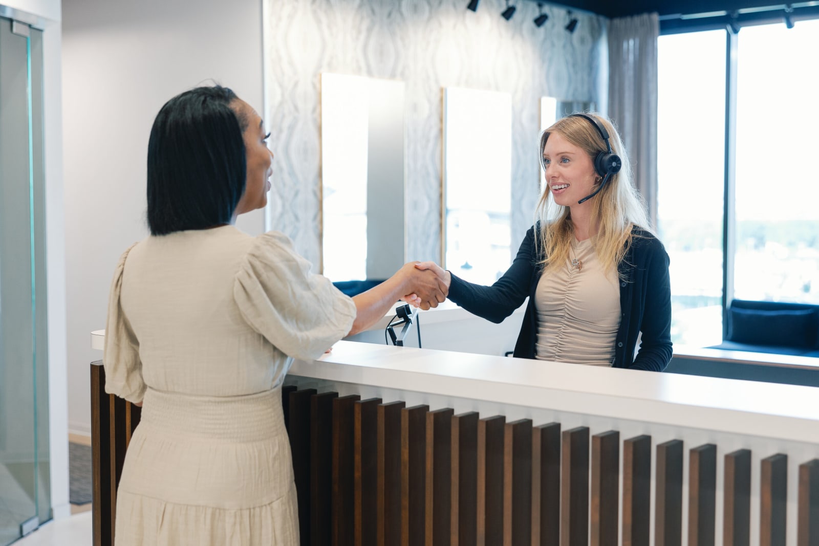 A Lucid Private Office receptionist wearing a headset smiling and shaking hands with a client over a white and wood-slatted front reception desk.